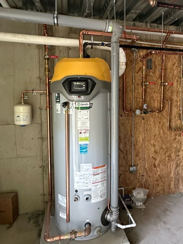 A gas-fired storage water heater with a yellow top cap and copper supply piping, a wall-mounted thermal expansion tank beside it, and a metal flue rising through the ceiling.
