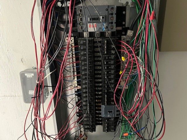 An open electrical panel with its cover removed, showing a main breaker, two rows of branch circuit breakers, and red, black, white, and green conductors landed on the bus and neutral bars.