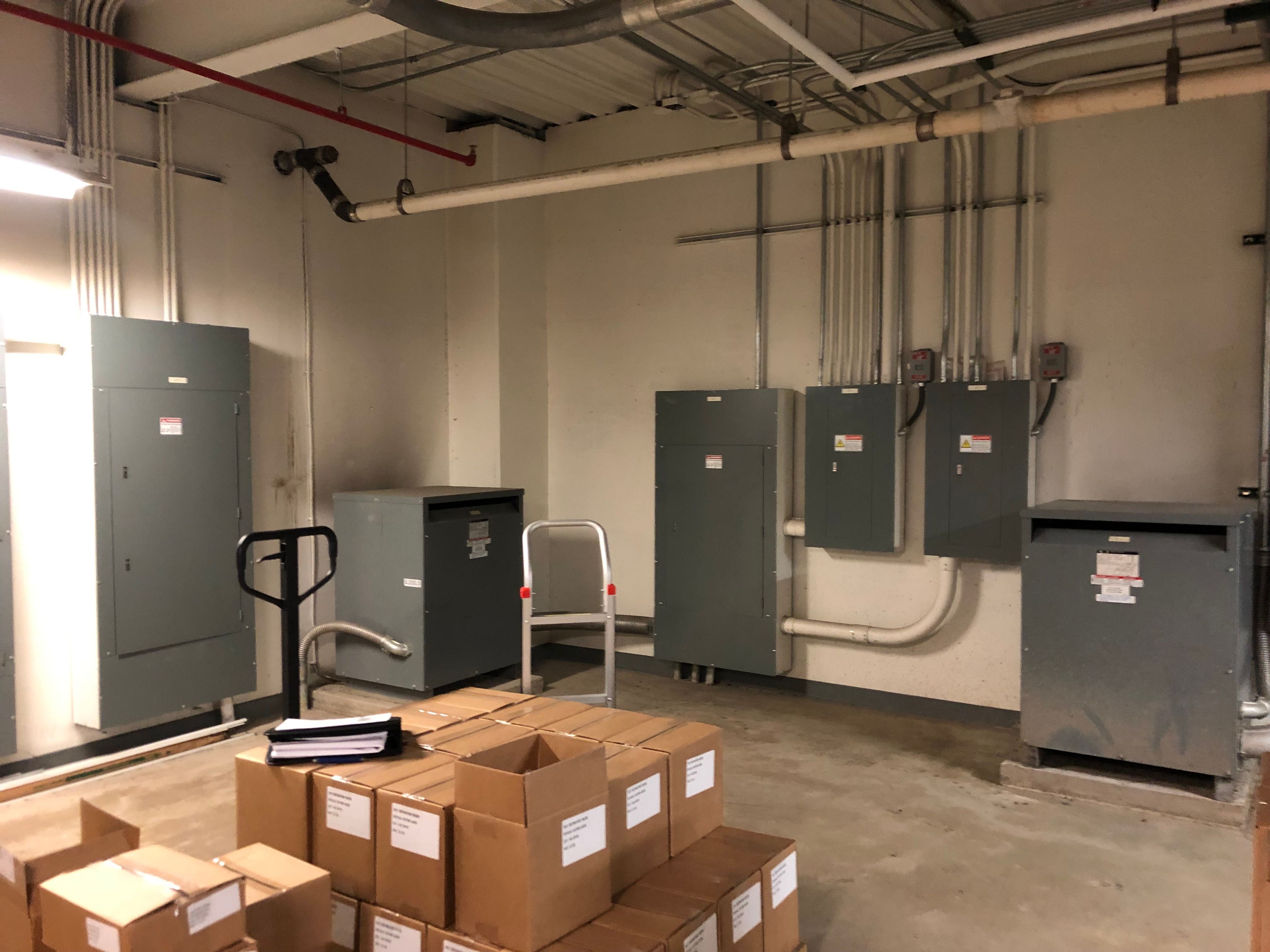 An electrical room with a large distribution panel, branch panelboards fed by conduit runs down the wall, and two floor-mounted dry-type transformers on concrete housekeeping pads.