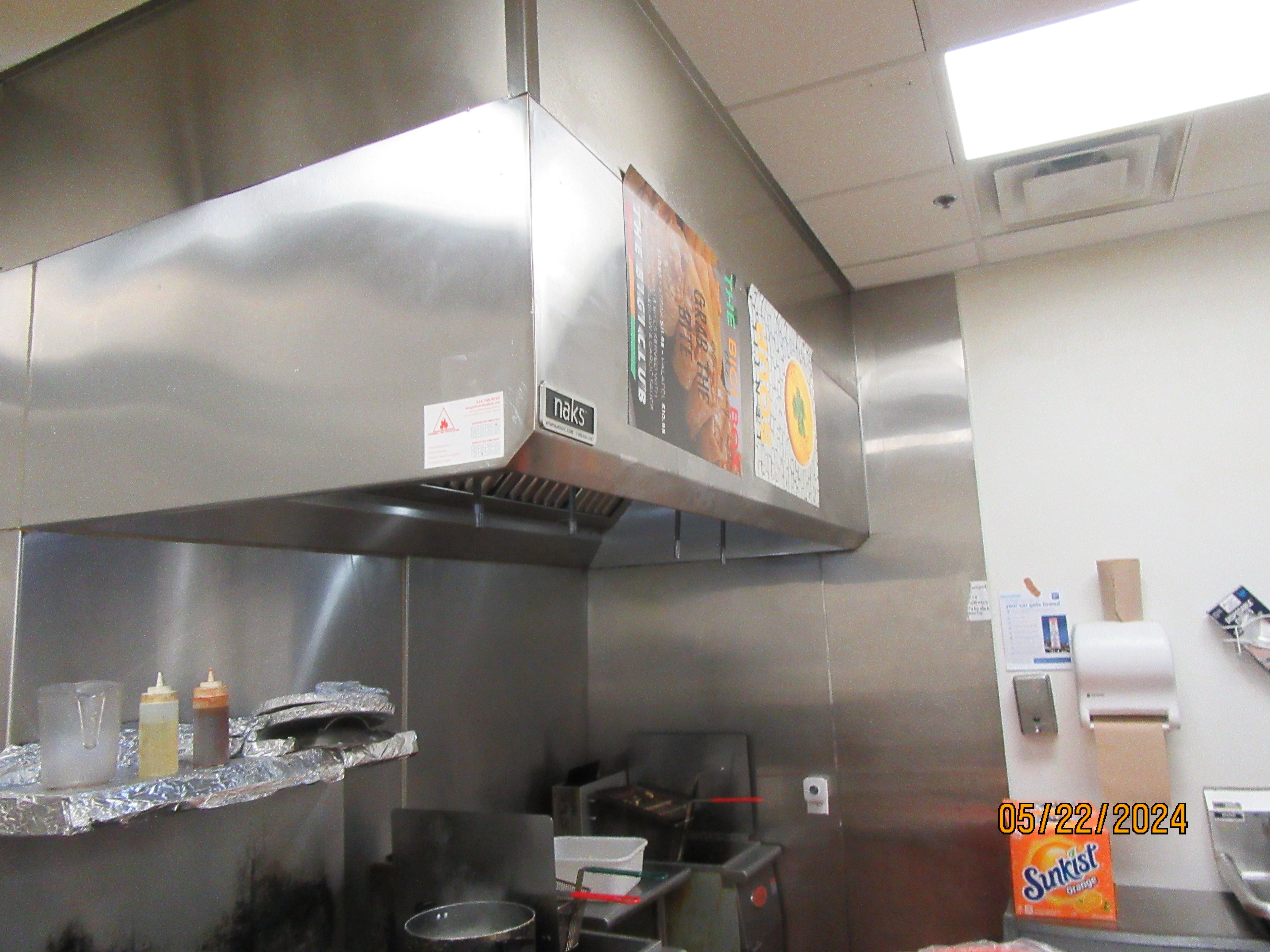 A stainless steel kitchen exhaust hood with grease filters and a fire-suppression nozzle, hung over a cooking line against a stainless wall panel.