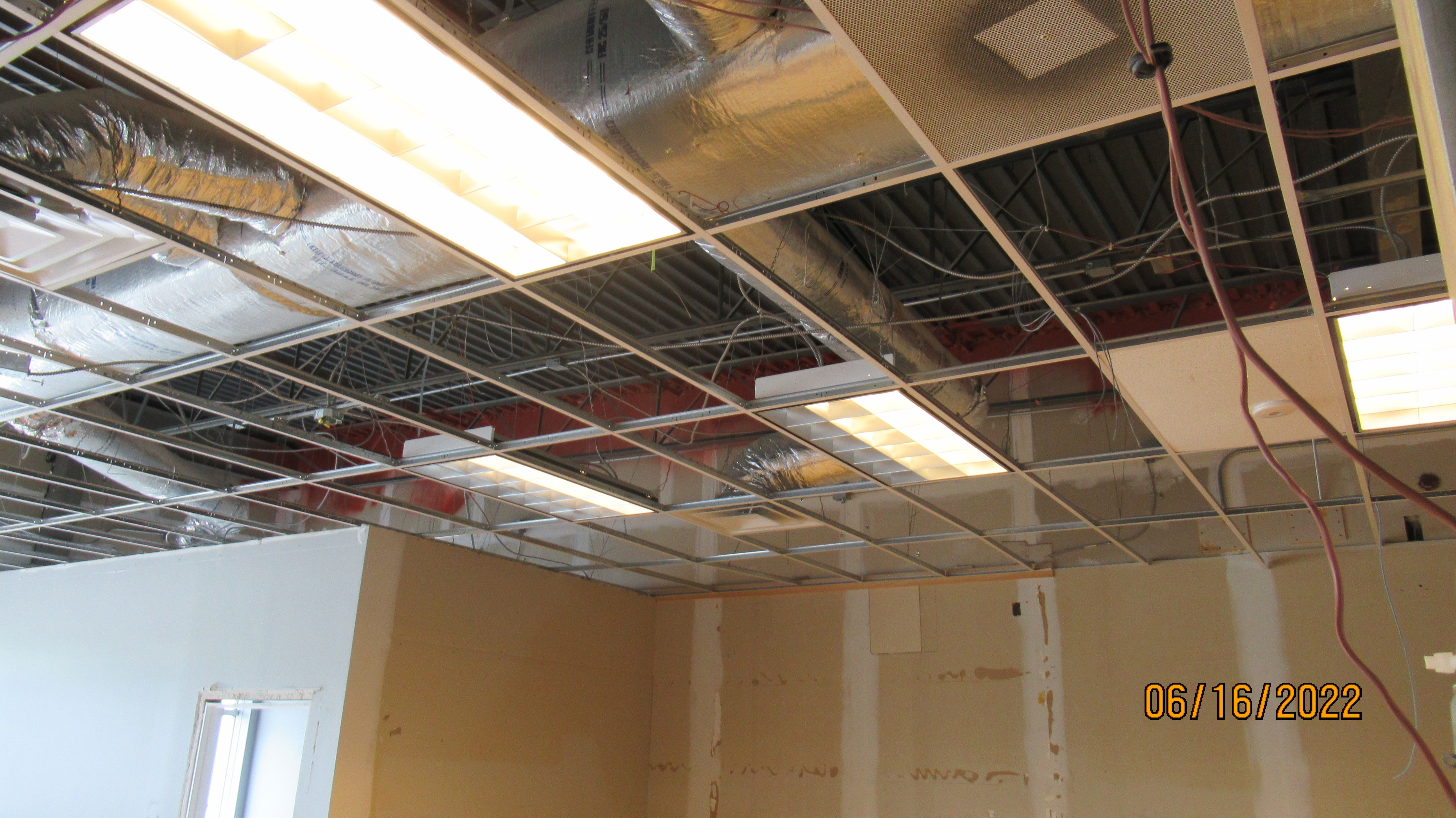 A dining room ceiling under construction, with recessed troffer light fixtures set into a suspended grid beside insulated flexible ductwork and armored cable runs above.