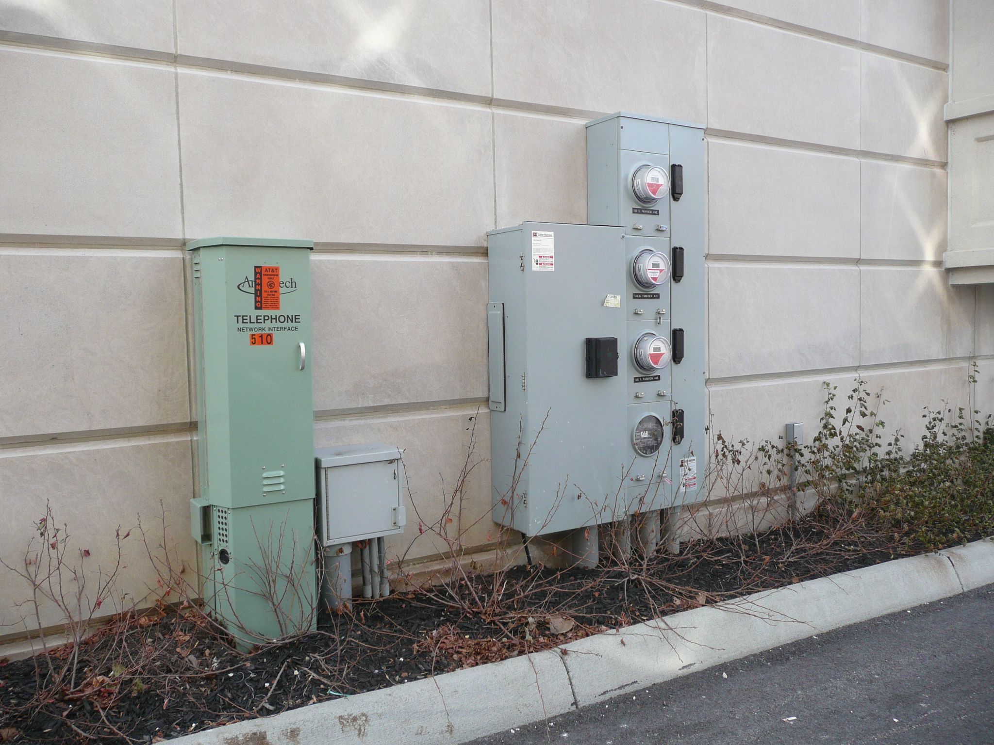 A multi-tenant electric meter center with four utility meters and their disconnects mounted on a precast concrete building wall, next to a green telephone network interface pedestal.