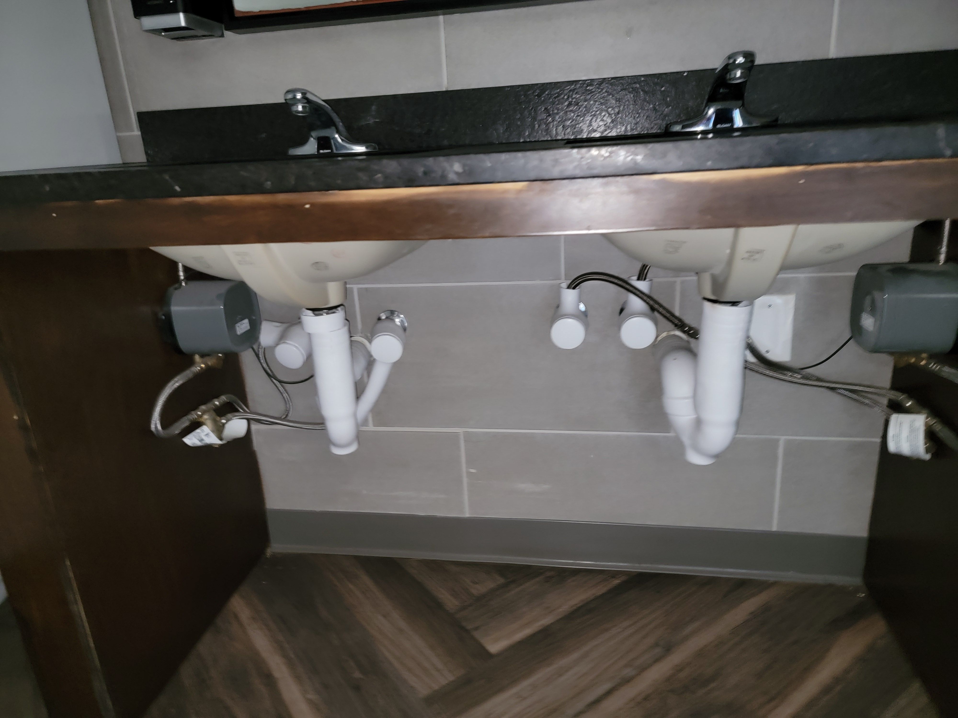 The underside of a restroom vanity showing two lavatory bowls with white P-traps, braided supply connections, angle stops, and the control boxes for their sensor faucets.