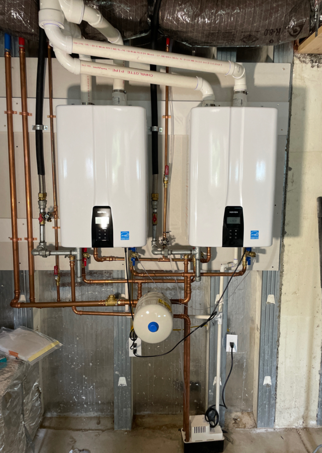Two wall-mounted tankless gas water heaters joined by a copper manifold with isolation valves, an expansion tank hanging below, and white PVC vent piping running up to the ceiling.