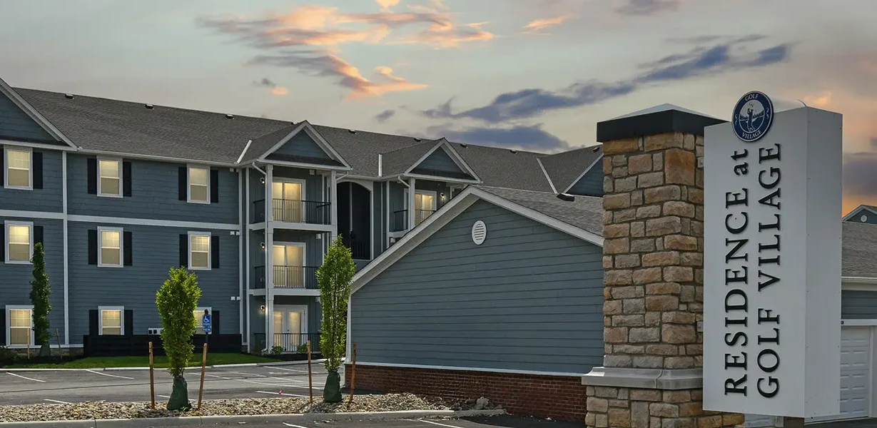 Three-story blue-gray apartment complex with stone entrance sign reading Residence at Golf Village at twilight.