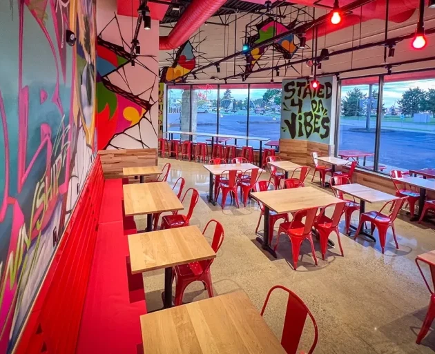 Colorful restaurant interior with red chairs, wooden tables, graffiti-style wall art, and exposed ductwork.